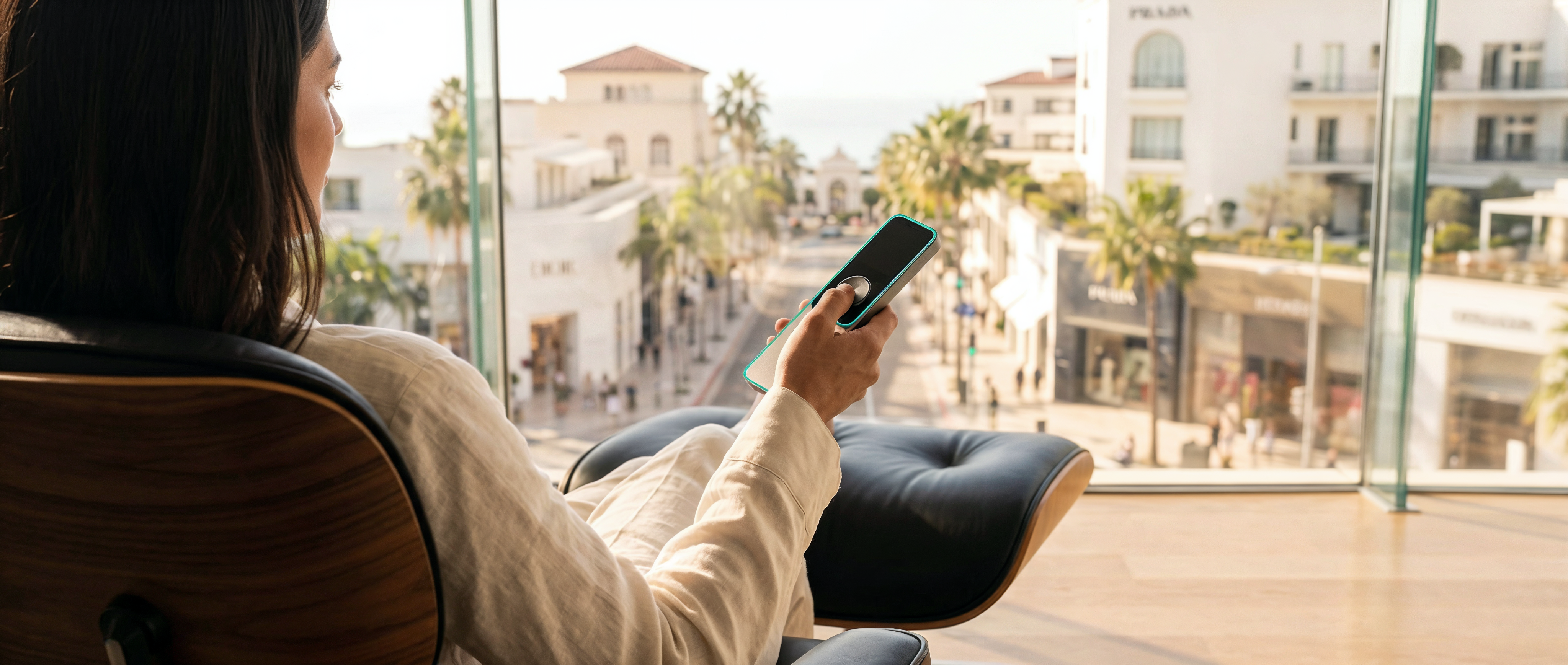 A wide-angle professional architectural photograph of a person sitting in a black leather Eames lounge chair, pressing a sleek Blicq smart remote button while looking through a panoramic window at a sunny Dubai retail street.